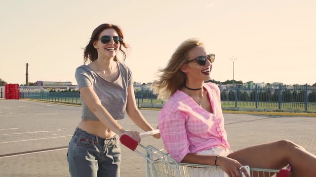 Fashion funny young hipster girls having fun at the shopping mall parking, roll each other in shopping cart on street. Slow motion