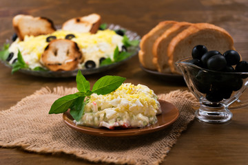 A portion of salad of sliced eggs, cheese, crab meat with mayonnaise, basil  leaves, pickled olives, toast, wooden background.