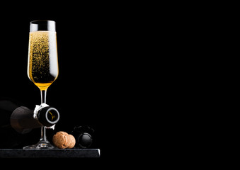 Elegant glass of yellow champagne with cork and wire cage and bottle on black marble board on black...