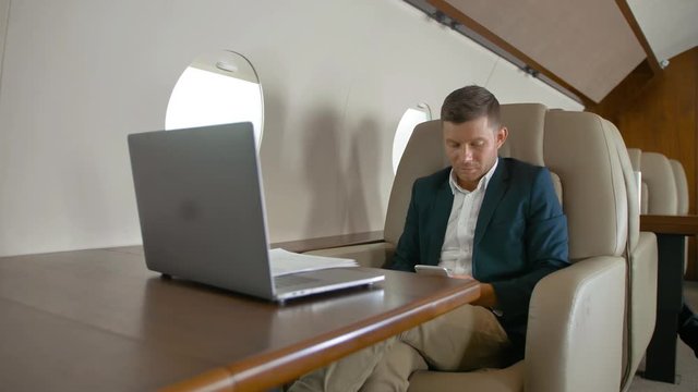 Handsome businessman chatting in modern smartphone in armchair in private jet cabin while plane flighting in the air. Luxury interior apartment look, slow motion.
