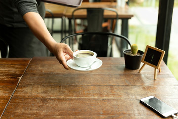 Waiter serving cup of coffee in with cell phone on cafe table.