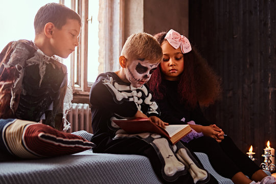 Three multiracial kids in scary costumes reading horror stories while sitting on bed in an old house.