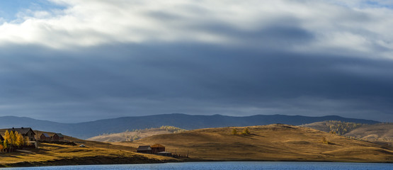 Wide panoramic photo of countryside landscape in autumn day with hill slopes illuminated by sunlight shining through the cloudy sky; meadows on sunset and long shadows of trees; yellow and blue shades