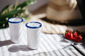 two white porcelain glasses, a hat, Lily of the valley bouquet, strawberries on a wooden table, sunlight on the terrace