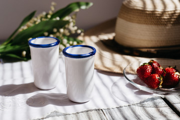 two white porcelain glasses, a hat, Lily of the valley bouquet, strawberries on a wooden table, sunlight on the terrace