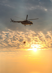 A fire fighter helicopter with a full basket of water flies against a beautiful sunset sky. Rescue operation. Forest Fire Prevention