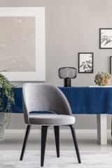 Modern chair at table with blue cloth in grey dining room interior with posters and lamp. Real photo