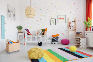 Poufs on colorful rug in scandi baby's bedroom interior with lamp, cradle and posters. Real photo