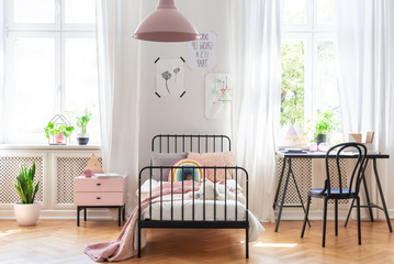 Chair at desk next to bed in pink and white girl's room interior with plants and windows. Real photo