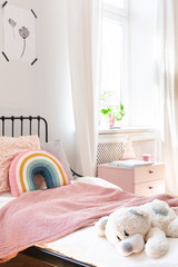 Plush toy and rainbow pillow on bed in white kid's bedroom interior with window. Real photo
