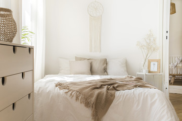 Blanket on white bed with pillows and wooden cabinet in minimal natural bedroom interior. Real photo - obrazy, fototapety, plakaty