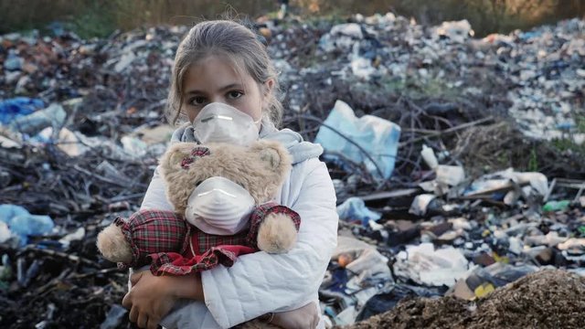 Pollution and Environmental Contamination. A young sad girl embracing a plush toy looks into the camera against the backdrop of a city dump. Wear respirators on the girl and on the toy.