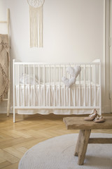 White cradle and shoes on wooden stool in baby's bedroom interior with round rug. Real photo