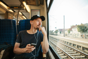 A young man listens to a music or podcast and looks out the window while the train is moving. - obrazy, fototapety, plakaty