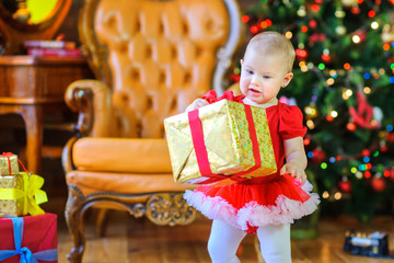 Fototapeta na wymiar little girl holding a big gift wrapped golden paper, on the background of a festive Christmas tree