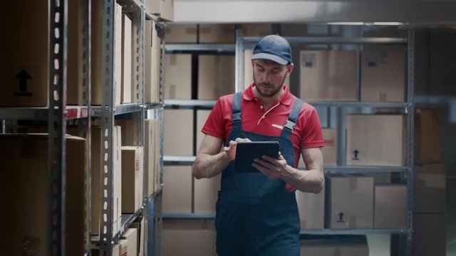 Warehouse Worker Uses Digital Tablet For Checking Stock, On the Shelves Standing Cardboard Boxes.