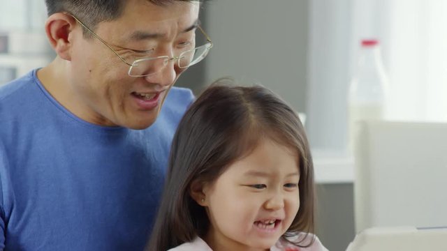 Slowmo medium shot of happy middle-aged Asian father in glasses laughing and talking to cute preschool-age girl sitting on his laps and trying to take bite from apple while watching cartoons on tablet