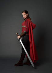  full length portrait of brunette girl wearing red medieval costume and cloak. standing pose  holding a sword on grey studio background.