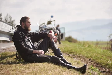 Tuinposter Athletic sportive motorcyclist in black leather clothing sitting at powerful motorbike on grassy roadside on blurred background of sunny distant hills under bright blue sky. © anatoliy_gleb