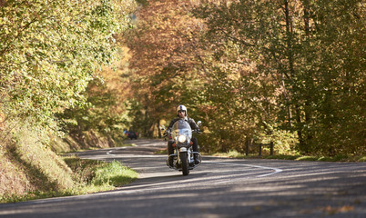 Bearded motorcyclist in helmet, sunglasses and black leather clothing riding motorbike along empty...