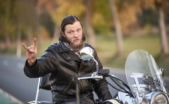 Young handsome bearded motorcyclist in black leather clothing sitting on shiny modern powerful motorbike showing horns sign on blurred vintage bokeh background of golden trees on bright sunny day.