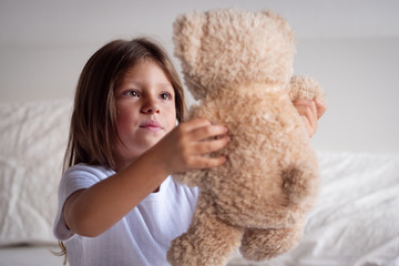 Portrait of a young girl while she is playing with her teddy bear on the bed.