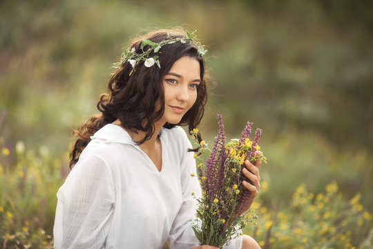 Beautiful young woman in white dress collecting wild flowers at the rural sunny landscape background in summer. Portrait of tender happy woman in wild field enjoying nature. Natural beauty model with