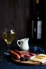 Charcuterie, grilled sausages, cheese, salami, plum olives and a glass of wine on a dark table