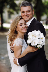 Groom in black tuxedo hugs tender stunning bride while they stand on the street of old European town
