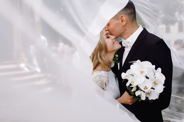 Groom in black tuxedo hugs tender stunning bride while they stand on the street of old European town