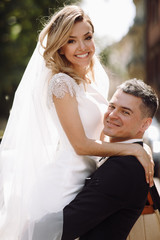 Groom in black tuxedo hugs tender stunning bride while they stand on the street of old European town