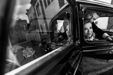 Look from the outside at adorable wedding couple in classy dresses sitting inside a black retro car and hugging each other tender