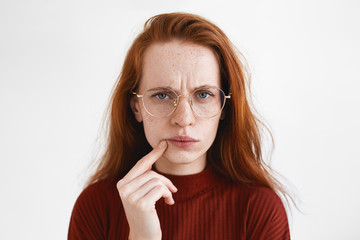 Picture of puzzled young red haired woman in spectacles frowning, having suspicious look, touching...
