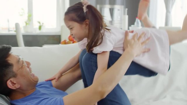 Handheld medium shot of Asian middle-aged father in glasses lying on sofa and lifting cute preschool-age girl with his legs