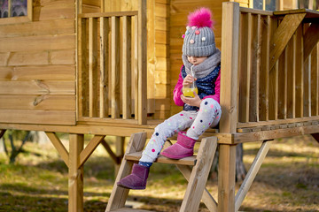 adorable little girl in a cap and jacket drinks lemonade in a house in an autumn forest