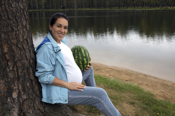 Attractive pregnant brunette with watermelon posing near the calm forest lake