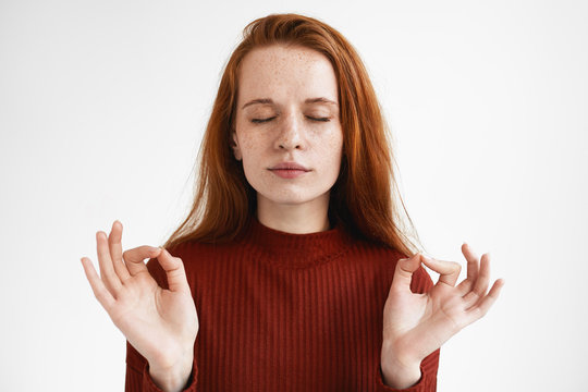 Zen, yoga and meditation concept. Picture of calm young red haired female in turtleneck keeping eyes closed and holding hands in mudra gesture, meditating after work, feeling relaxed and peaceful