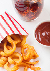 Curly fries fast food snack in paper container with glass of cola and ketchup on stone kitchen background. Unhealthy junk food