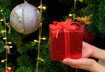 Female hands holding a red glitter square shaped gift box with silver sequined ball ornament, and Christmas decorations in background 
