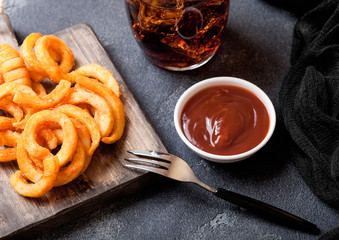 Curly fries fast food snack on wooden board with ketchup and glass of cola on stone kitchen background. Unhealthy junk food
