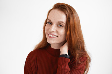 Obraz premium Dreamy pretty girl looking at camera with joyful smile, adjusting her loose red hair. Attractive young European female model in maroon turtleneck advertising shampoo or hair care pproduct, smiling