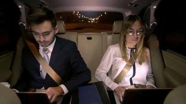 View from driver seat of young cheerful businesswoman video calling on laptop and businessman using laptop and drinking coffee during ride in the backseat of corporate car in the night