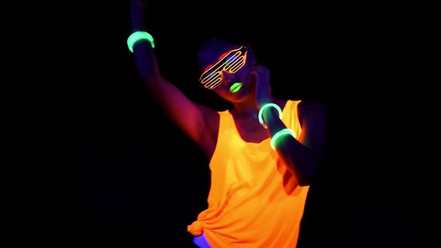 Beautiful sexy woman with UV face paint, glowing clothing, glowing glasses, bracelet dancing in front of camera, half body shot. Asian woman. Party concept.
