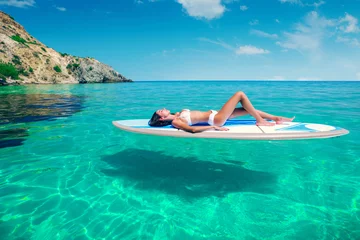 Rolgordijnen Jonge mooie vrouw ontspannen in de zee op een SUP-bord. Het meisje zonnebaadt op het strand van het eiland op vakantie. © kuznetsov_konsta