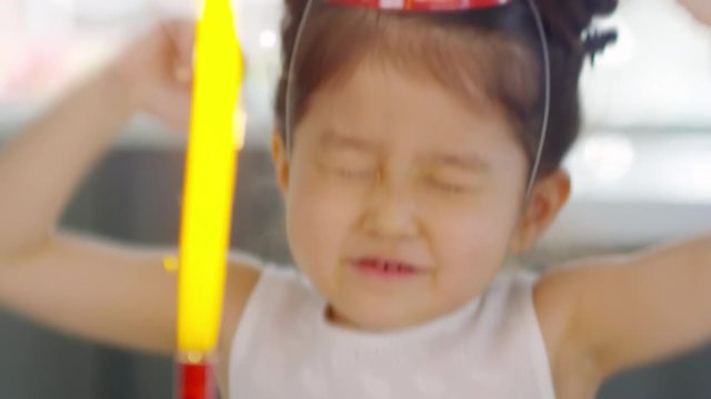 Dolly shot of adorable Asian girl in party hat celebrating before birthdate cake with firework candle