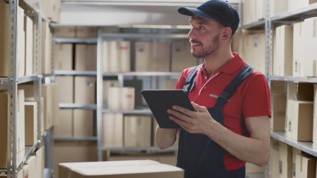 Warehouse Worker Uses Digital Tablet For Checking Stock, On the Shelves Standing Cardboard Boxes, Packages.
