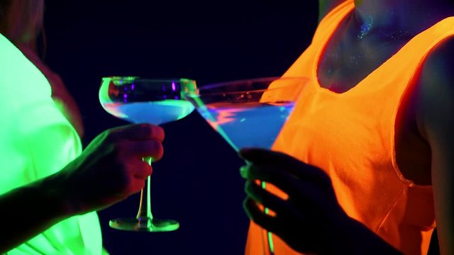 Beautiful sexy women with UV face paint, glowing bracelets, drinks, glowing clothing dancing together in front of camera, Close up of drinks. Caucasian and asian woman. Party concept.