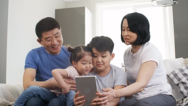 Tracking shot of happy Asian parents and children sitting on sofa and playing with tablet: they are laughing and taking selfies together