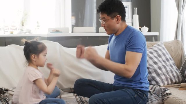 Tracking shot of cheerful Asian middle-aged father in glasses and cute little girl sitting on sofa and playing rock-paper-scissors