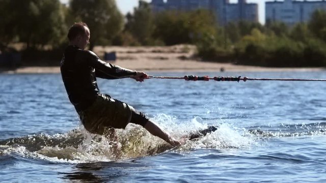 Wakesurfer riding surf board on waves in sunny day. Extreme water sport in slow motion. Msn riding on water. Wake surfing sportsman training